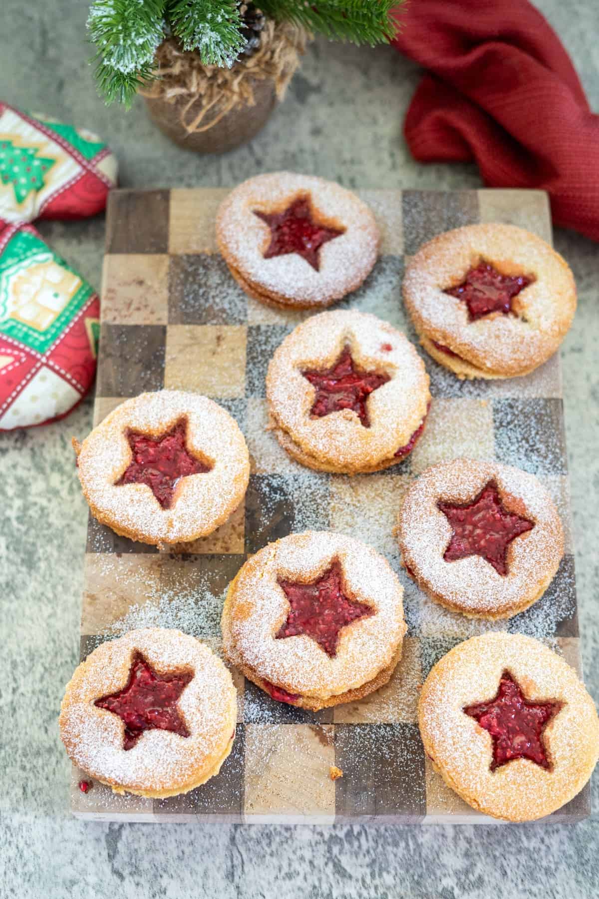 Seven star-shaped almond flour linzer cookies, filled with jam and dusted with powdered sugar, rest on a checkered wooden board alongside festive oven mitts and a small potted evergreen.