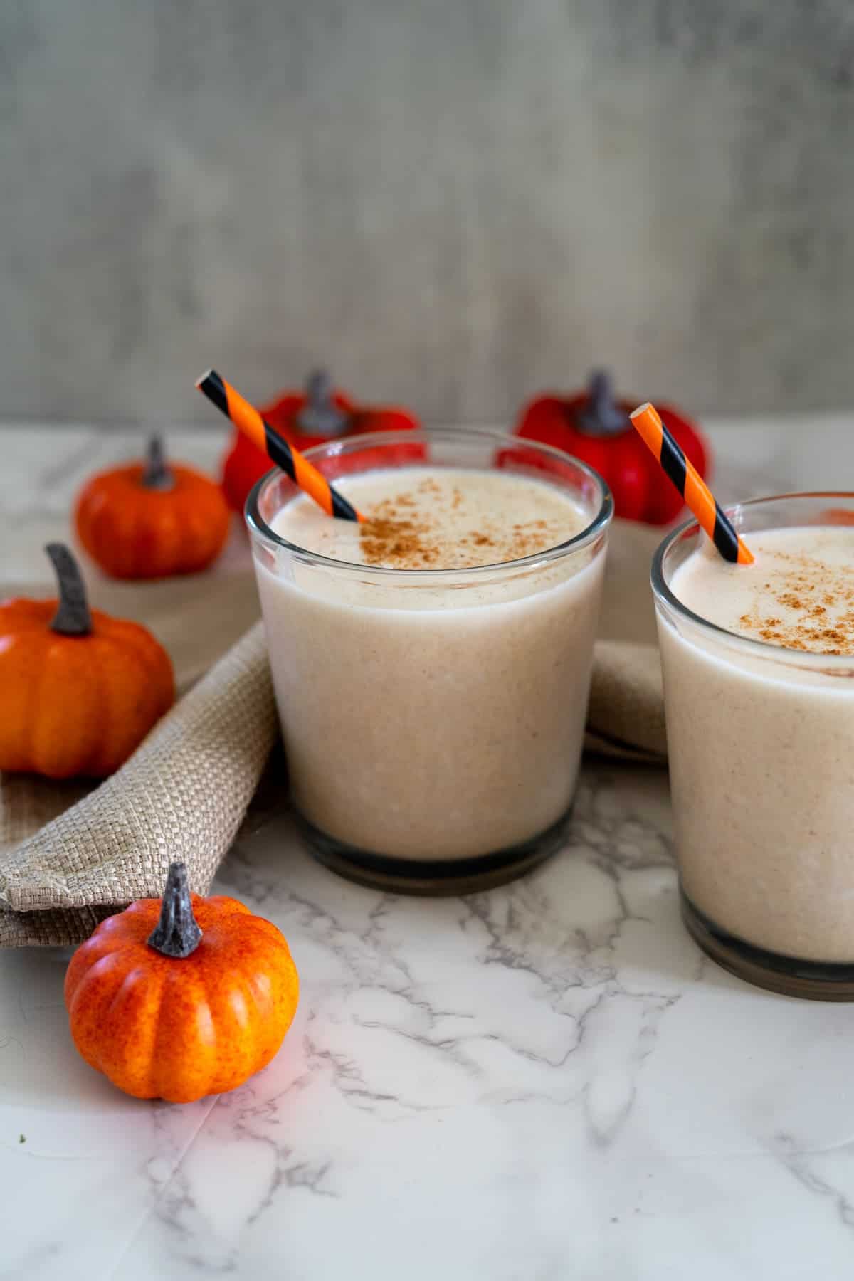 Two glasses of frothy pumpkin spice drinks with cinnamon, surrounded by small decorative pumpkins and a burlap cloth on a marble surface.