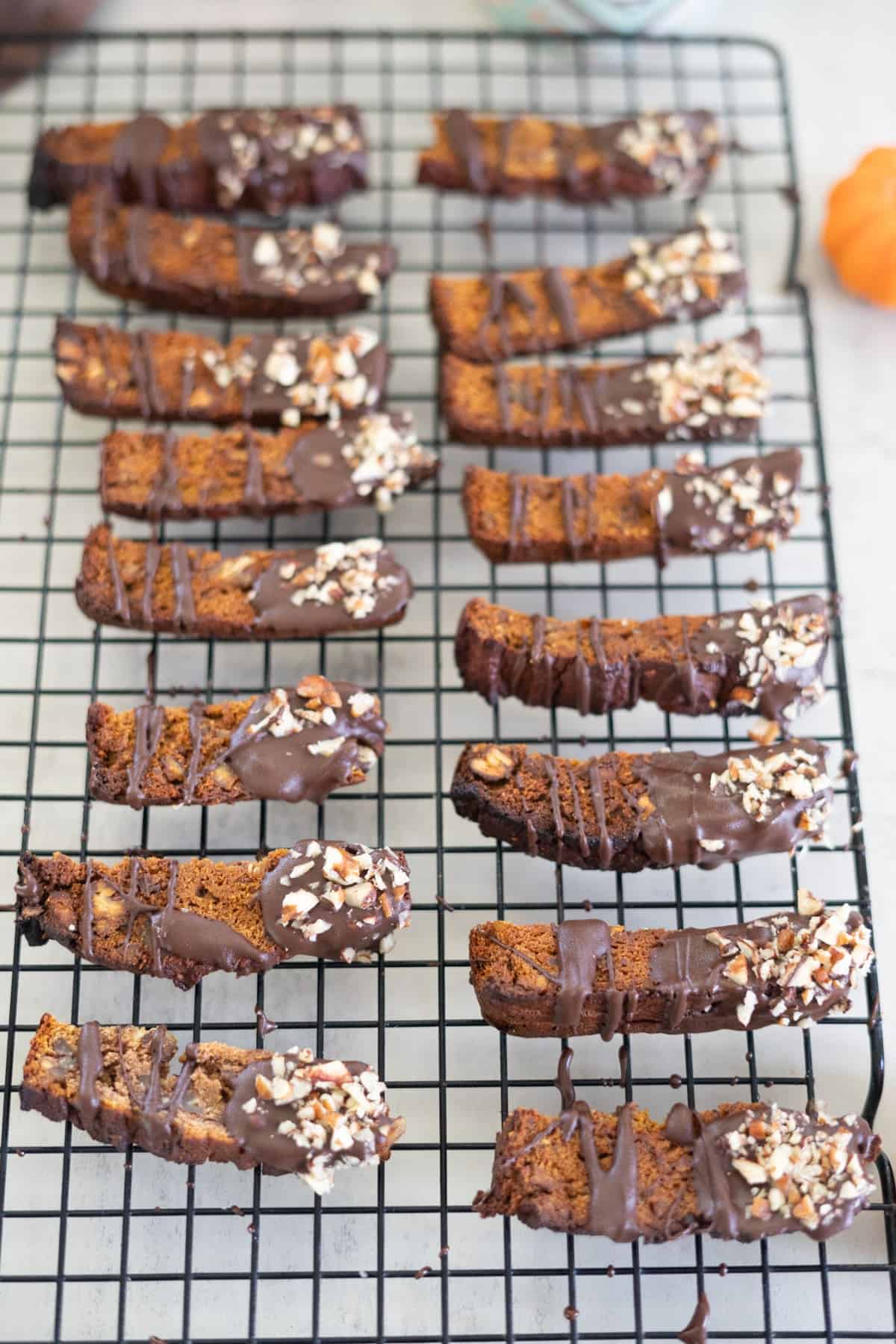 Pumpkin biscotti dipped in chocolate with chopped nuts rest on a cooling rack.