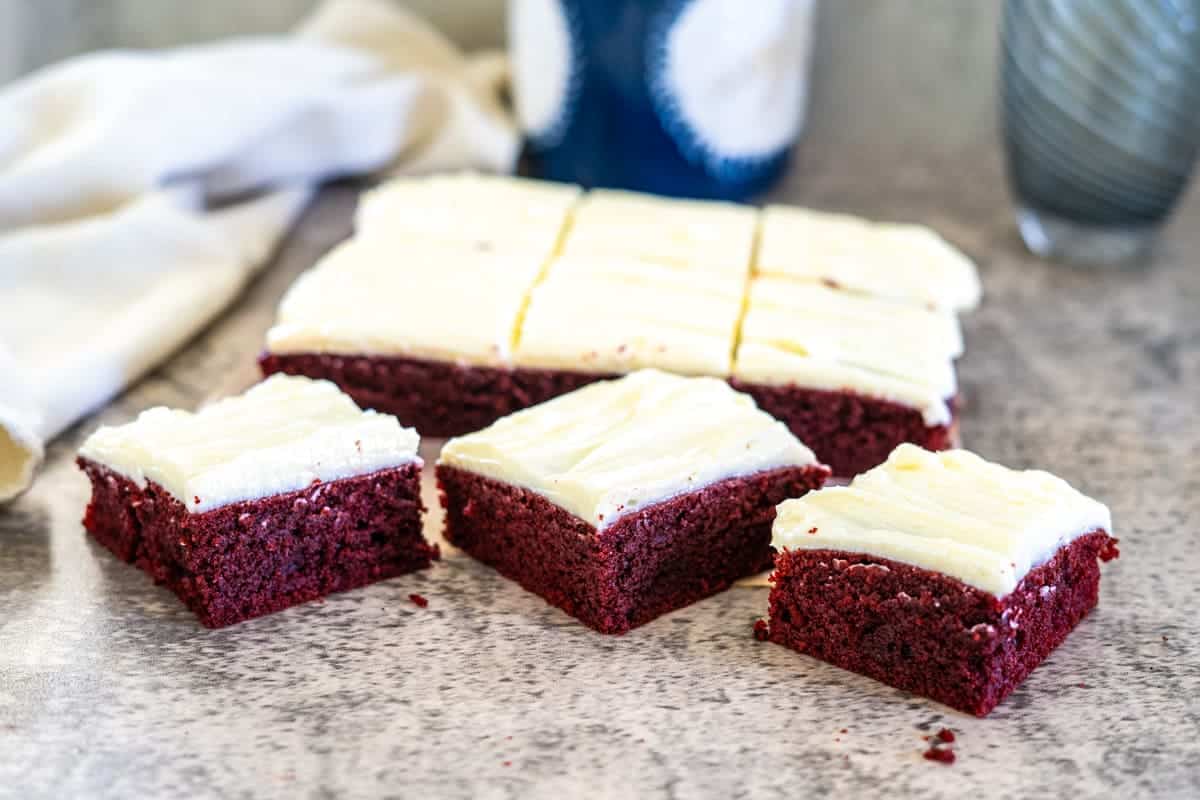 Three square pieces of red velvet cake with cream cheese frosting sit in the foreground, while a larger rectangular piece rests on the marble counter behind them, reminiscent of indulgent keto red velvet brownies.