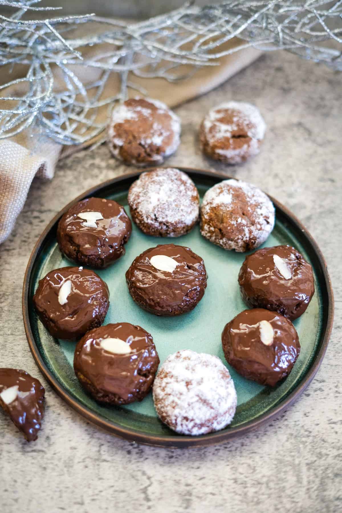 A plate of chocolate-covered cookies and powdered sugar cookies, including delectable low carb lebkuchen, is displayed on a grey surface with decorative silver netting in the background.