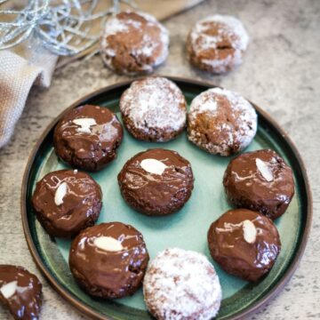 A plate of chocolate cookies topped with almonds and powdered sugar, arranged on a gray countertop.