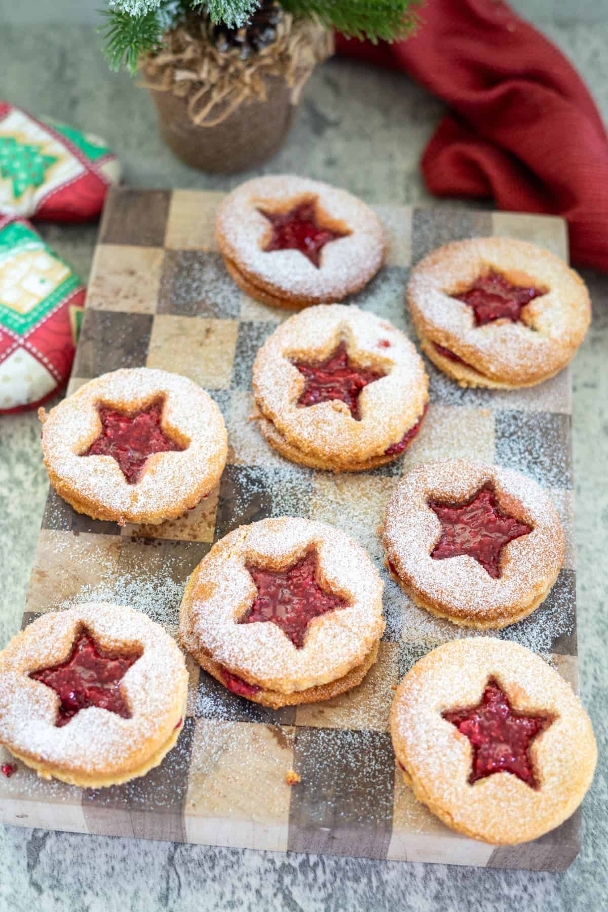 A wooden board showcases eight almond flour Linzer cookies, each filled with red jam and topped with powdered sugar, featuring star-shaped cutouts. A small potted plant and red cloth gracefully accent the background.