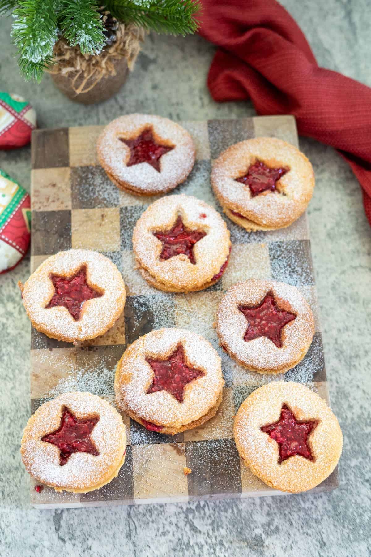 Cookies with star-shaped cutouts filled with red jam are arranged on a checkered wooden board, dusted with powdered sugar. A red cloth and decorative ornaments are nearby.