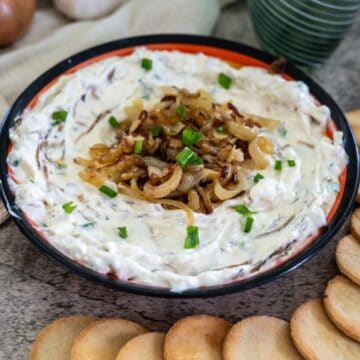 A bowl of creamy dip topped with caramelized onions and chopped green herbs, surrounded by round crackers on a wooden surface.