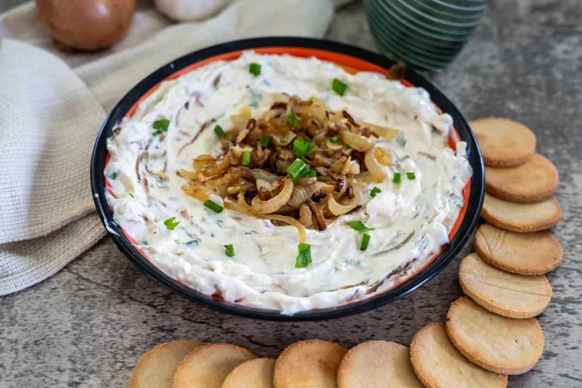 A bowl of creamy dip topped with caramelized onions and green onions, surrounded by round crackers on a gray surface.