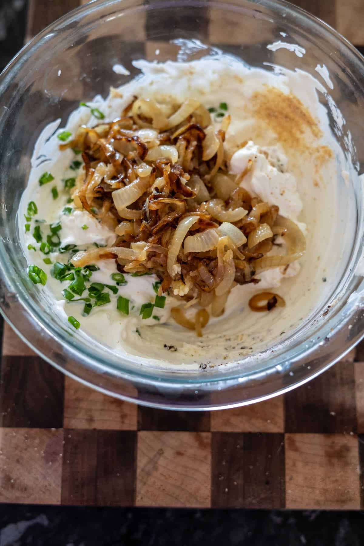 Glass bowl with sour cream, caramelized onions, chopped chives, and seasoning on a wooden cutting board.