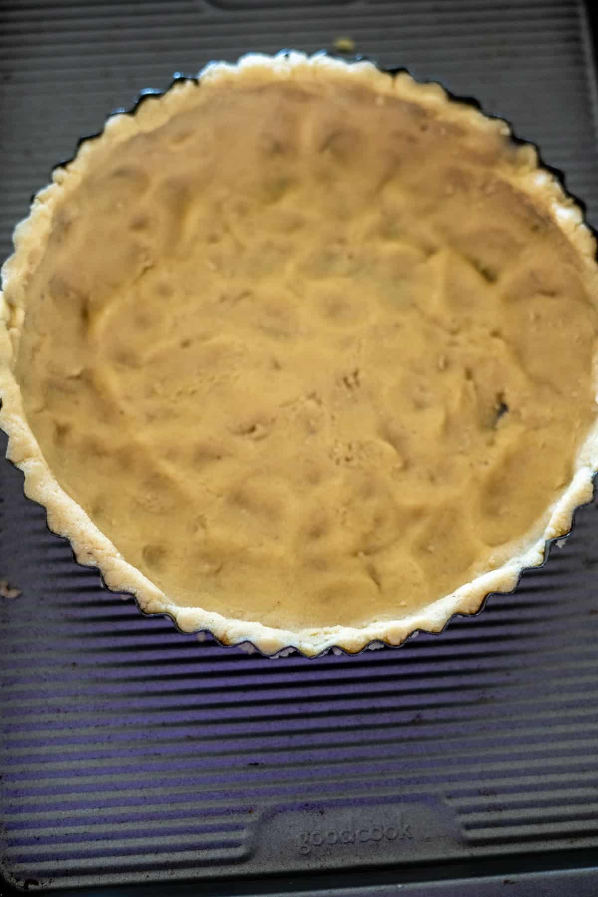 A keto chocolate tart sits unbaked, its beige batter nestled in a fluted pie crust on a ribbed baking sheet.