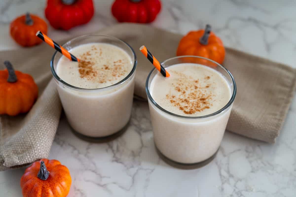 Two glasses of creamy pumpkin spice protein shake with orange-striped straws, topped with cinnamon, set on a marble surface with small decorative pumpkins.