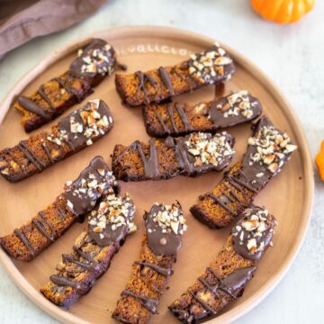 Chocolate-dipped biscotti topped with chopped nuts arranged on a round wooden plate.