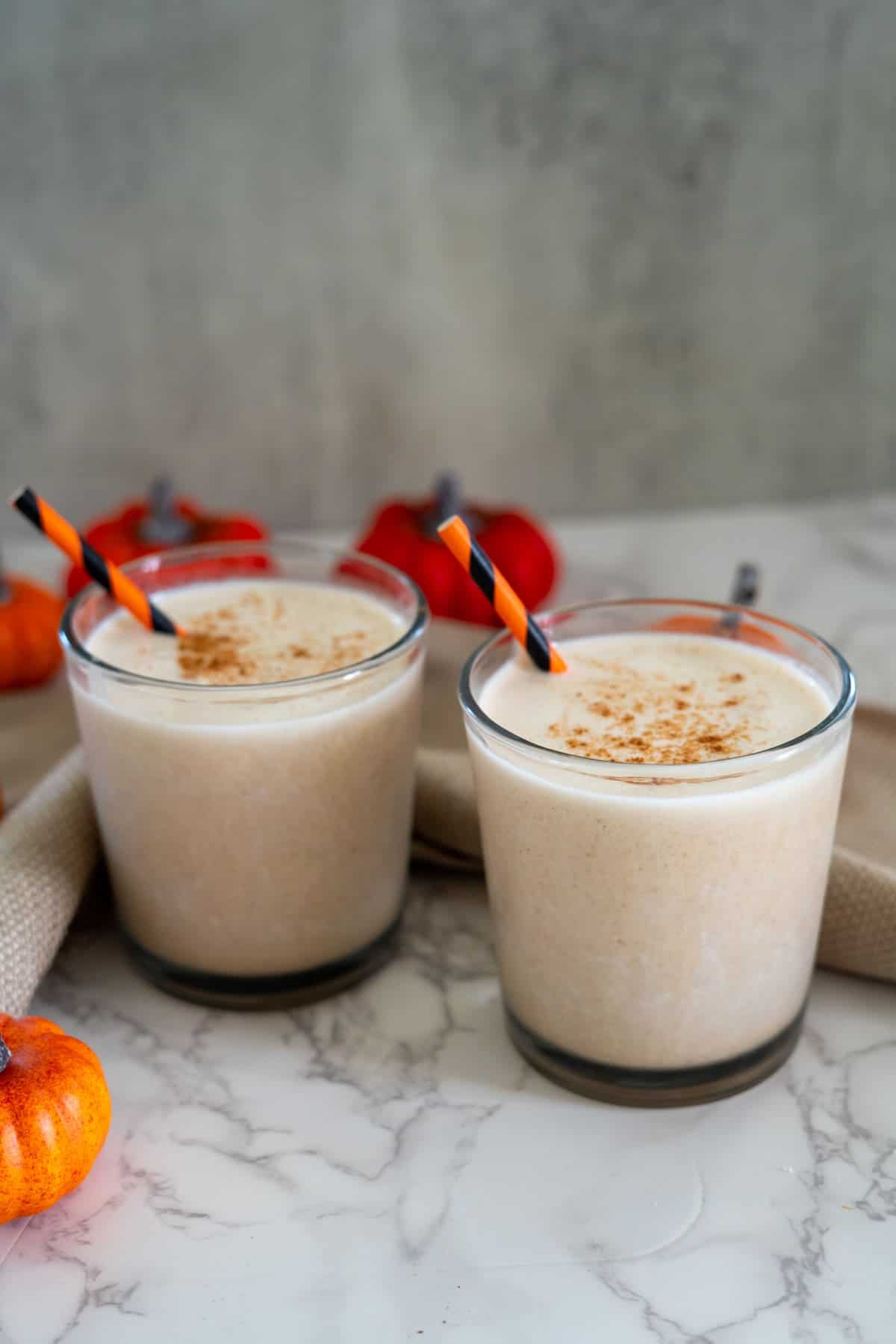 Two glasses of pumpkin spice smoothies with striped straws, topped with cinnamon. Small pumpkins are placed around the glasses on a marble surface.