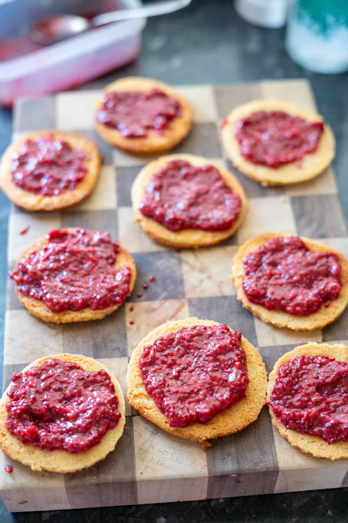 Nine almond flour Linzer cookies spread with red berry jam lay on a checkered wooden cutting board.