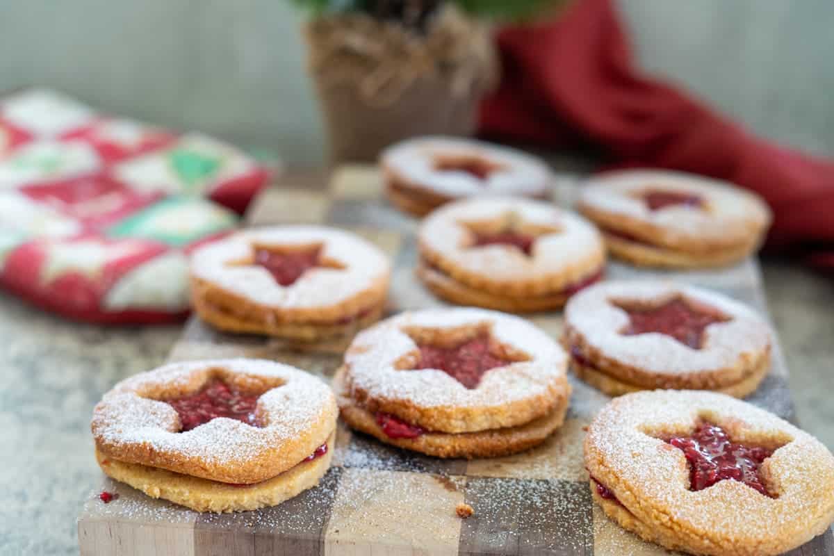 Nine star-shaped almond flour linzer cookies, dusted with powdered sugar and filled with raspberry jam, are arranged on a wooden board. A red cloth and a potted plant add charm in the background.