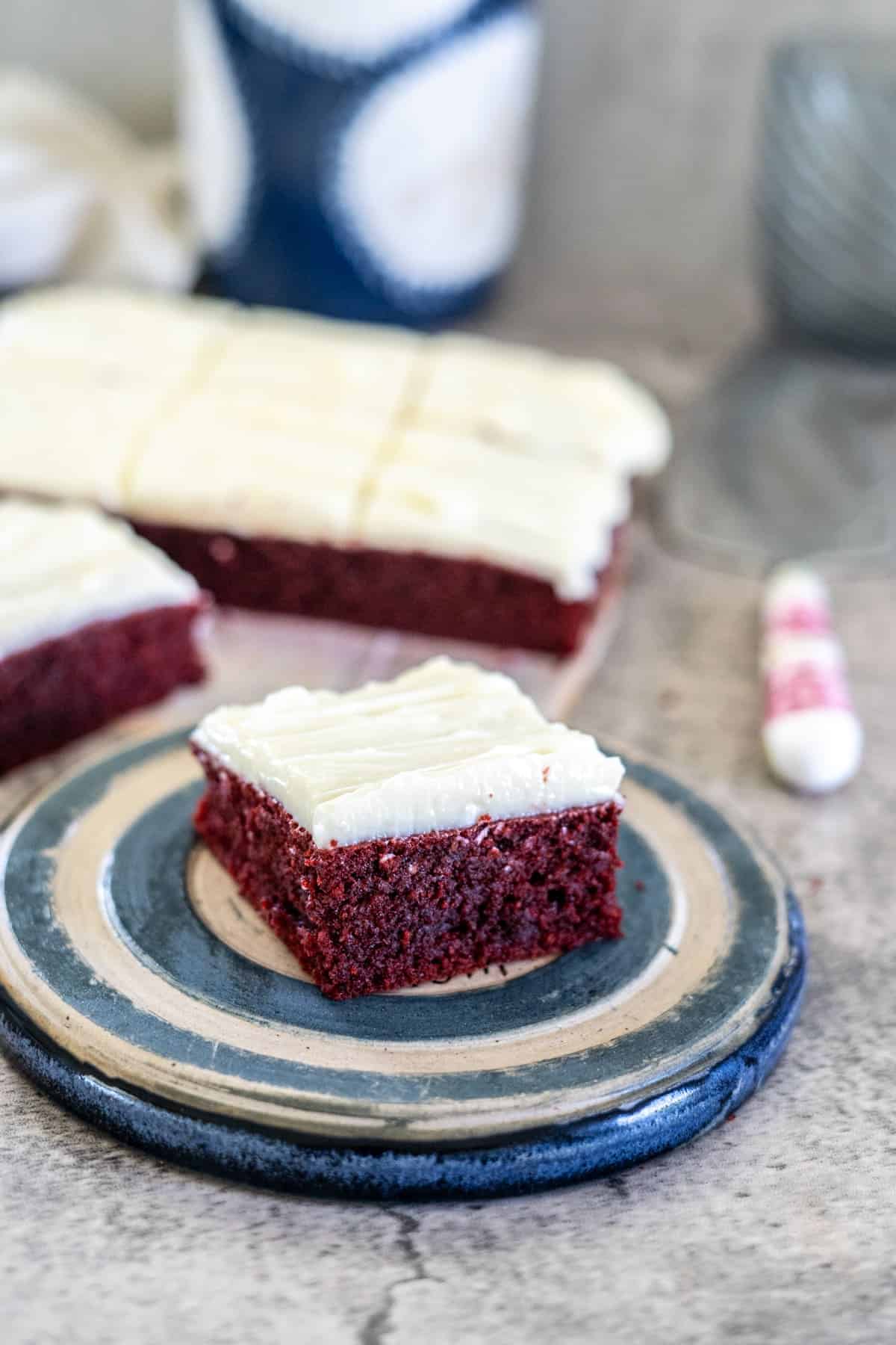 A square piece of frosted red velvet cake sits on a ceramic plate, with more cake pieces and a blurred background.