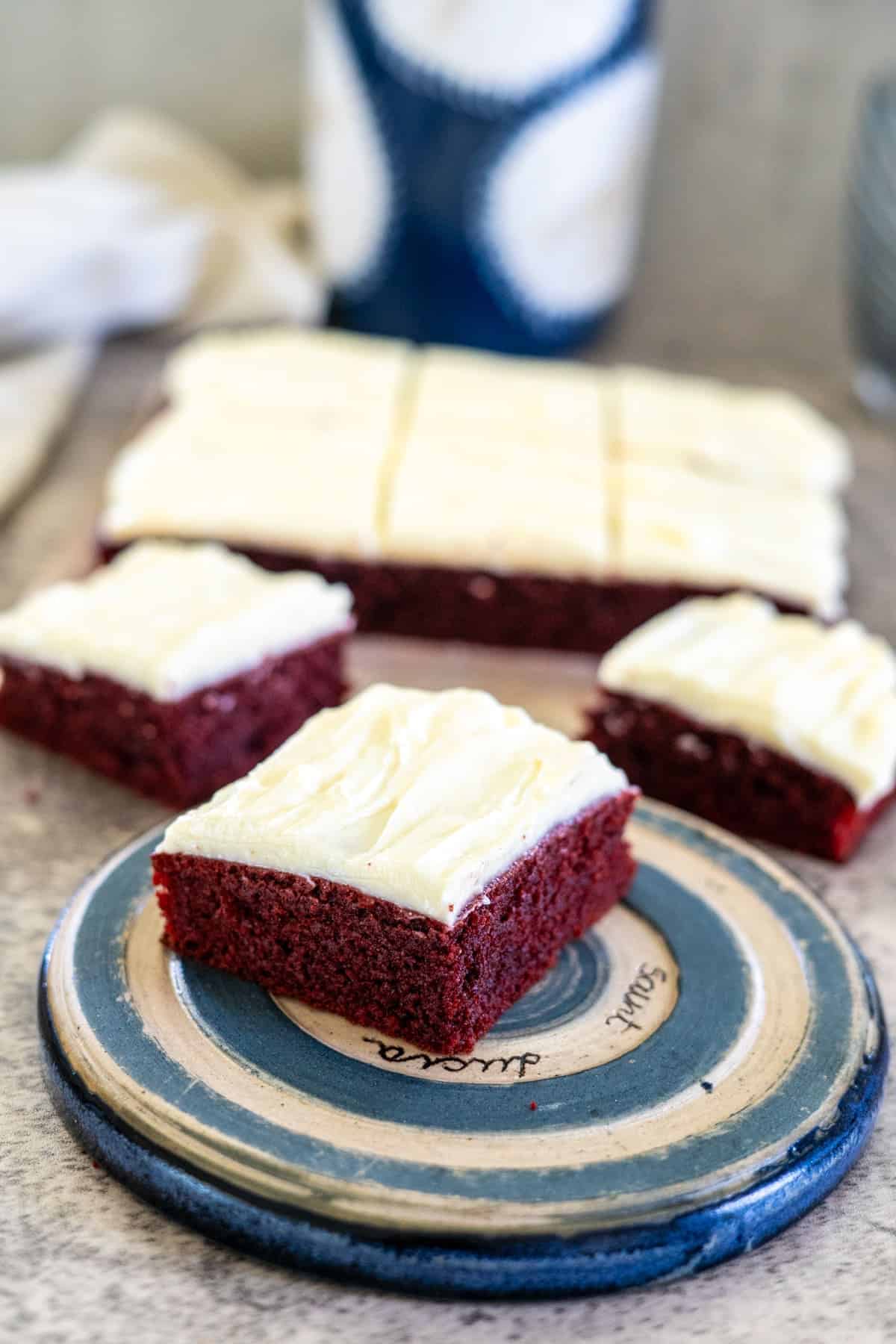 A slice of red velvet cake with white frosting on a plate, reminiscent of indulgent keto red velvet brownies, with more delectable pieces in the background.