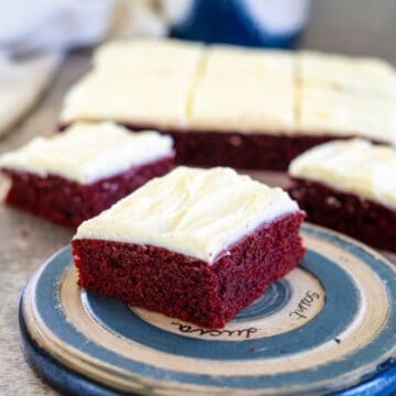 A slice of red velvet cake with cream cheese frosting is on a round plate. The full cake and other slices are in the background.