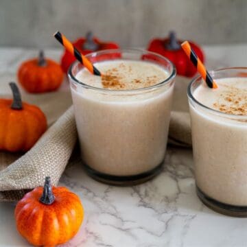 Two glasses of pumpkin spice smoothies with striped straws on a marble surface, surrounded by mini pumpkins and a beige cloth.