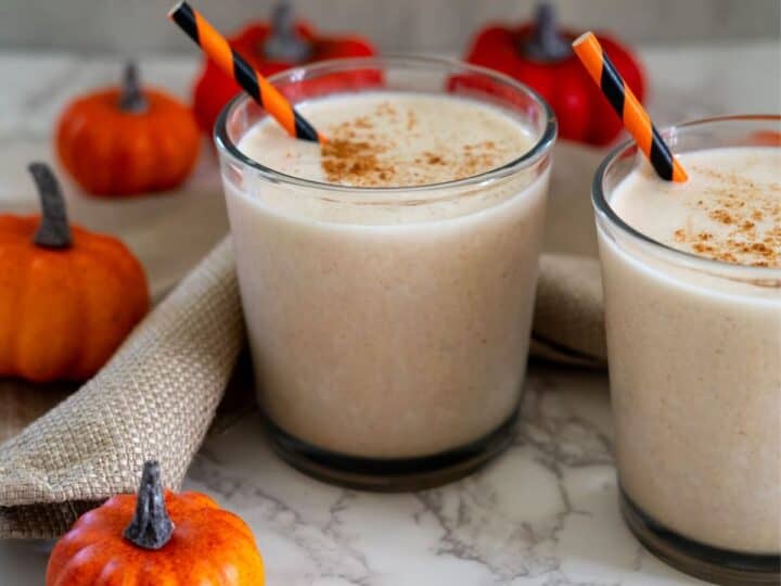 Two glasses of pumpkin spice smoothies with striped straws on a marble surface, surrounded by mini pumpkins and a beige cloth.