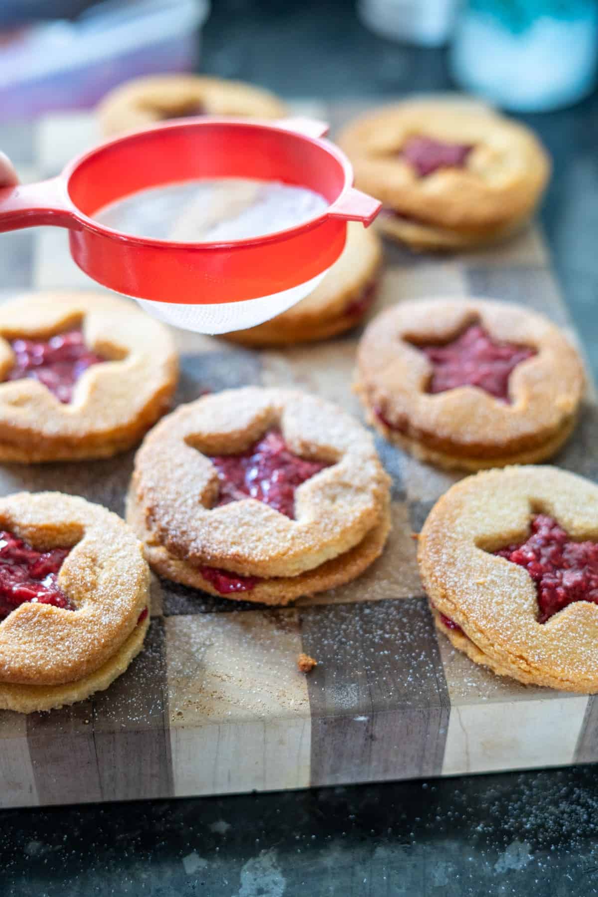 Person dusting sugar over almond flour linzer cookies, with jam-filled star-shaped cutouts, on a wooden board.