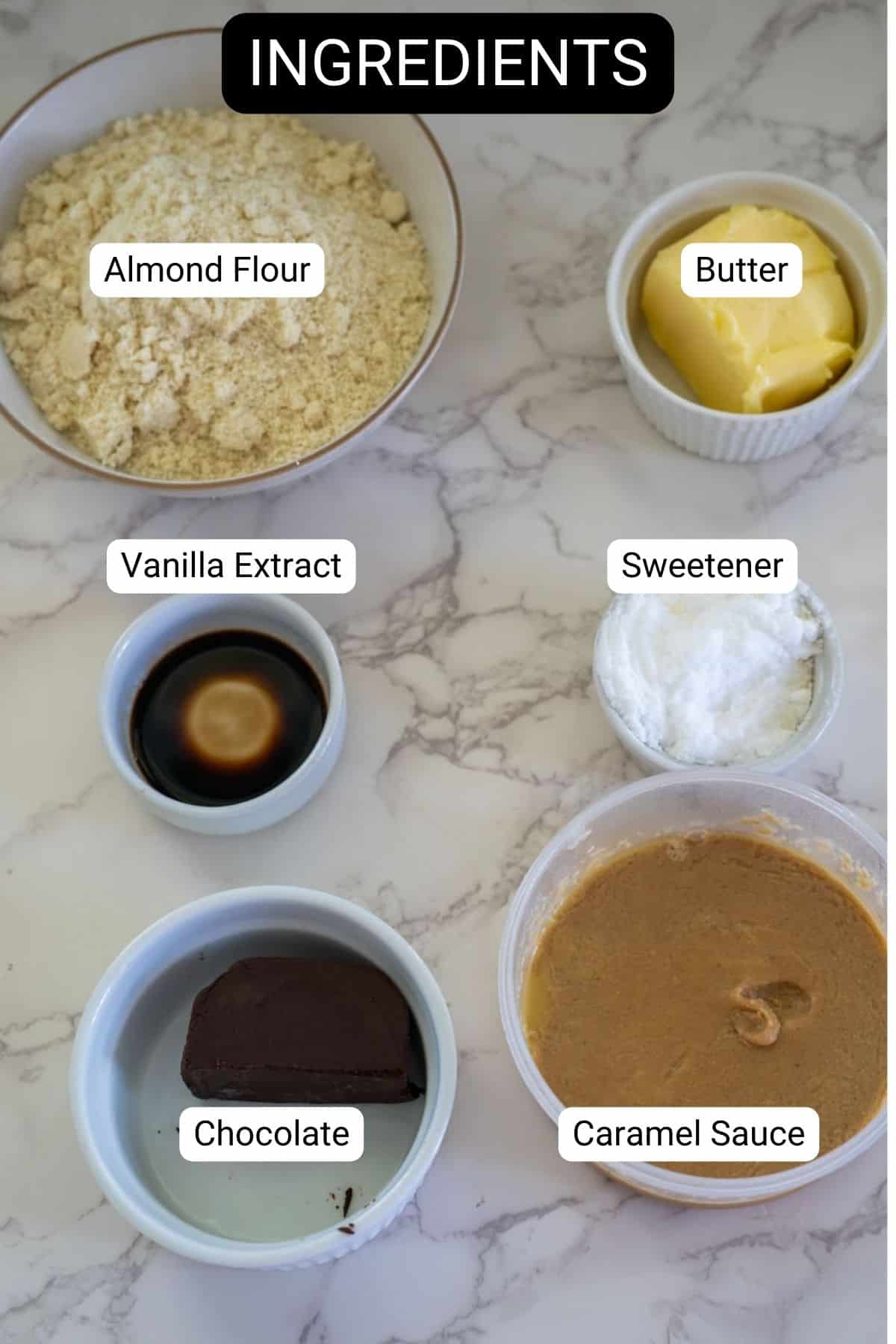 A marble countertop displays labeled bowls of almond flour, butter, vanilla extract, sweetener, chocolate, and caramel sauce.