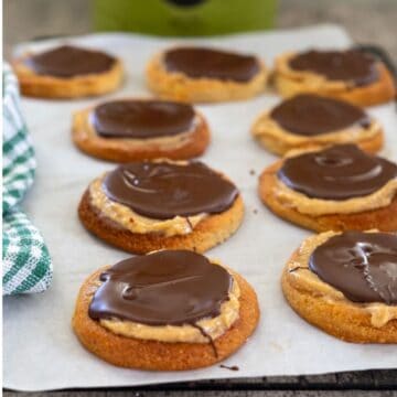 Cookies topped with peanut butter and chocolate are arranged on parchment paper. A green and white cloth is partially visible on the left.