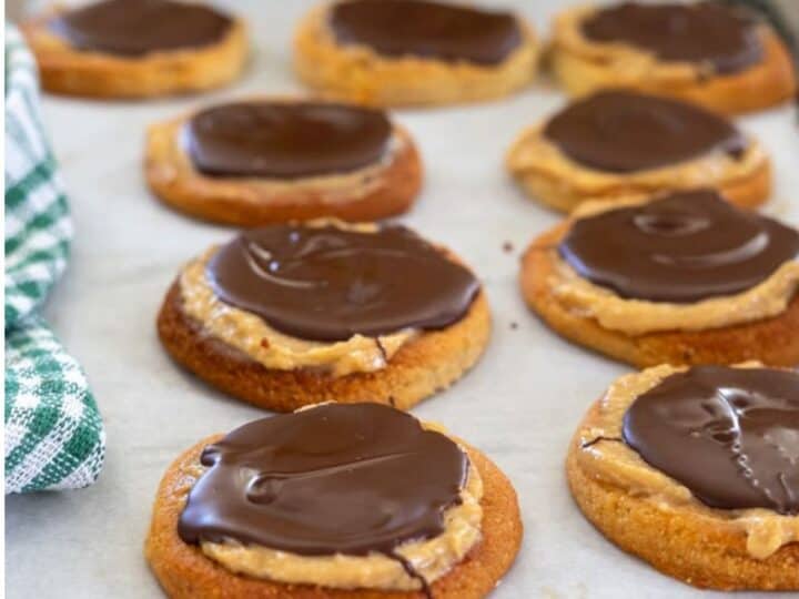 Cookies topped with peanut butter and chocolate are arranged on parchment paper. A green and white cloth is partially visible on the left.