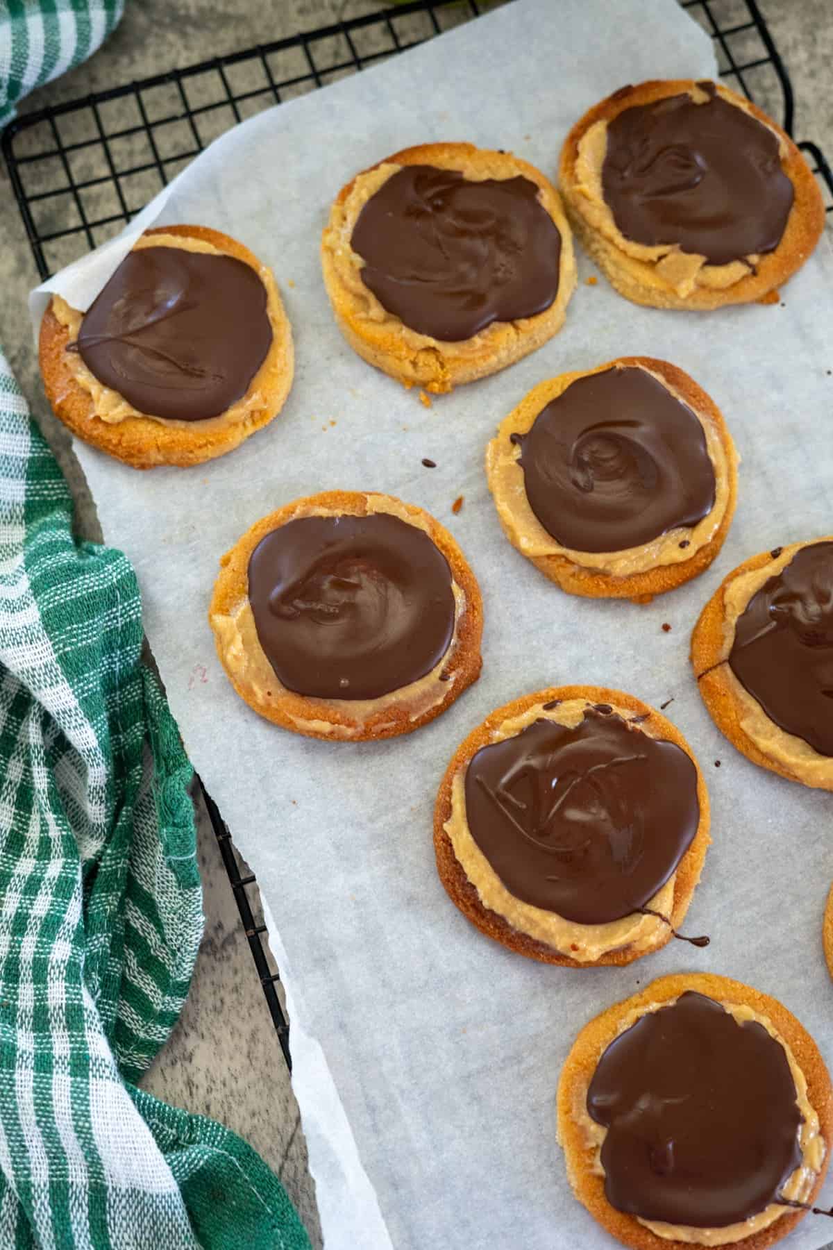 Keto Twix cookies with chocolate coating on top are arranged on parchment paper over a cooling rack. A green and white checkered cloth is partially visible on the side, adding a touch of rustic charm.