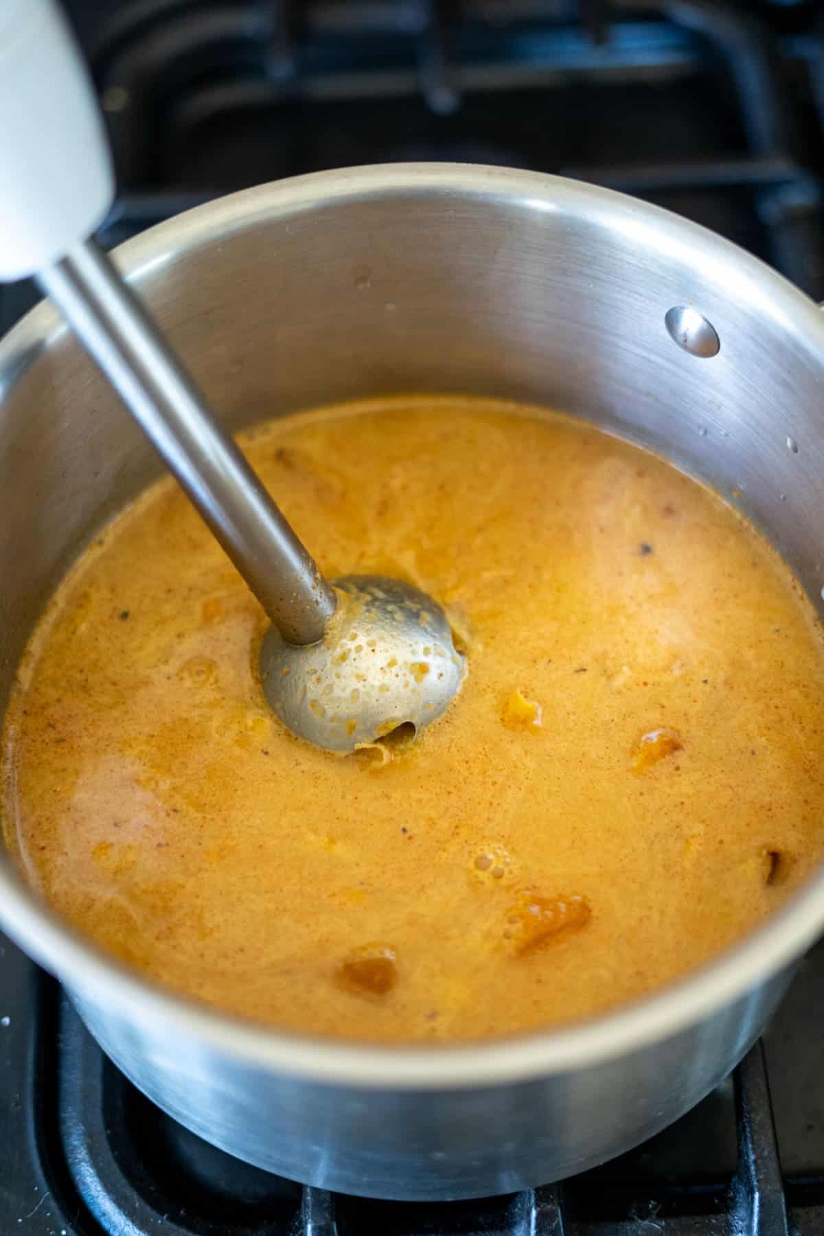 Immersion blender smoothly blending a hearty butternut squash and carrot soup with chunks in a metal pot on the stove.