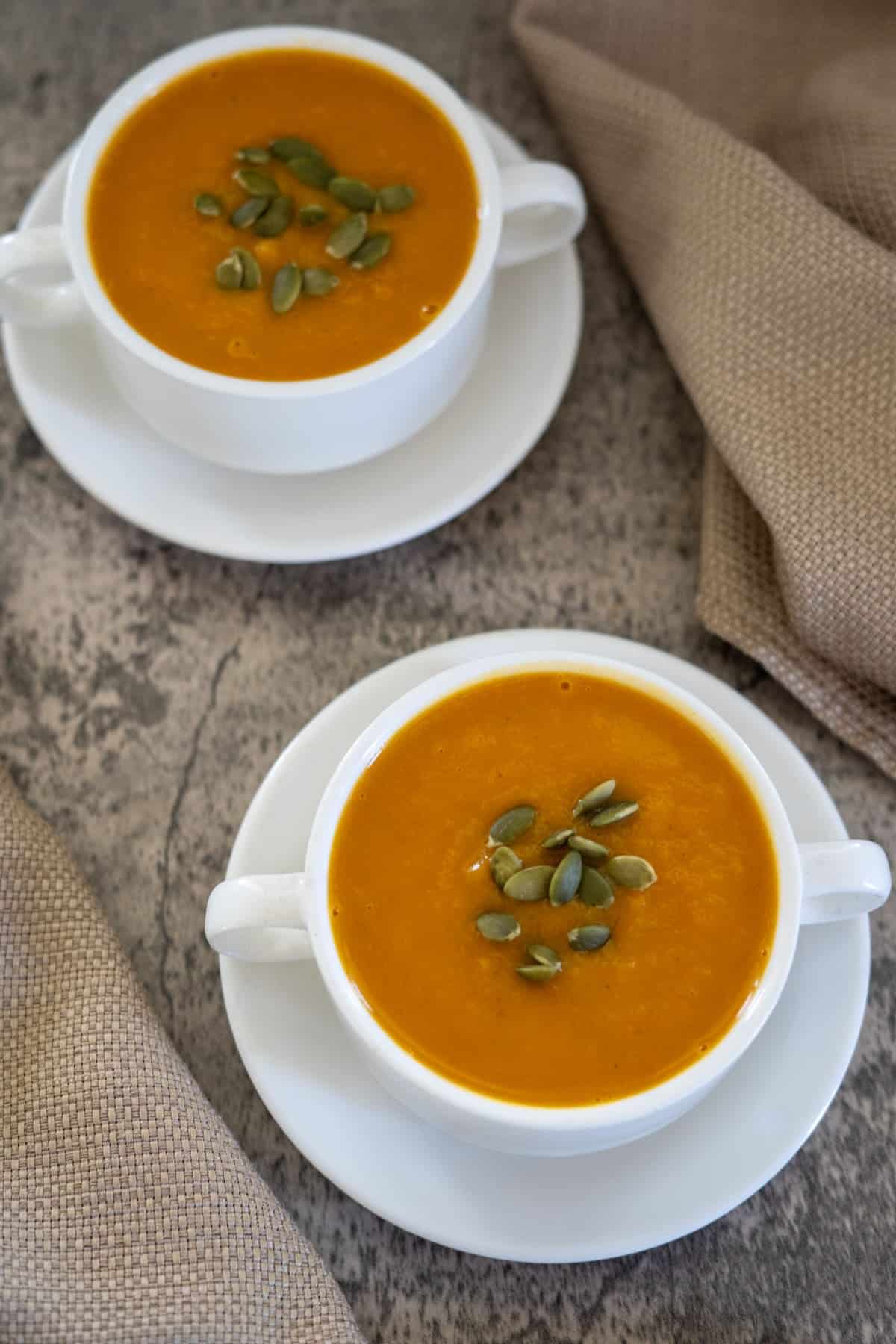 Two white bowls of butternut squash and carrot soup topped with pumpkin seeds rest on a stone surface, accompanied by beige cloth napkins.