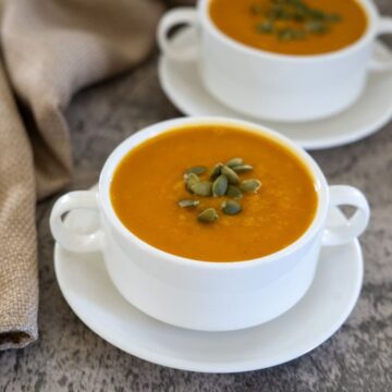 Two white bowls of pumpkin soup topped with seeds, placed on matching saucers on a gray textured surface, with a beige napkin nearby.