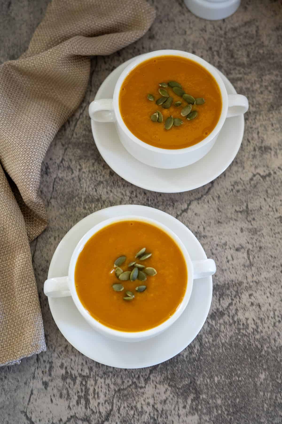 Two white bowls of pumpkin soup topped with seeds on white saucers, set on a textured gray surface with a beige cloth beside them.