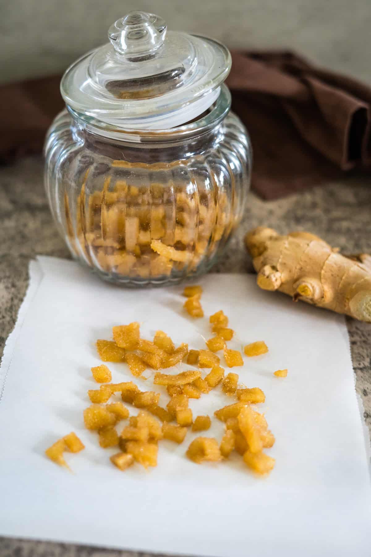 A glass jar filled with crystalized ginger sits on a countertop with some pieces spread out on white paper. A piece of fresh ginger is placed beside the jar.