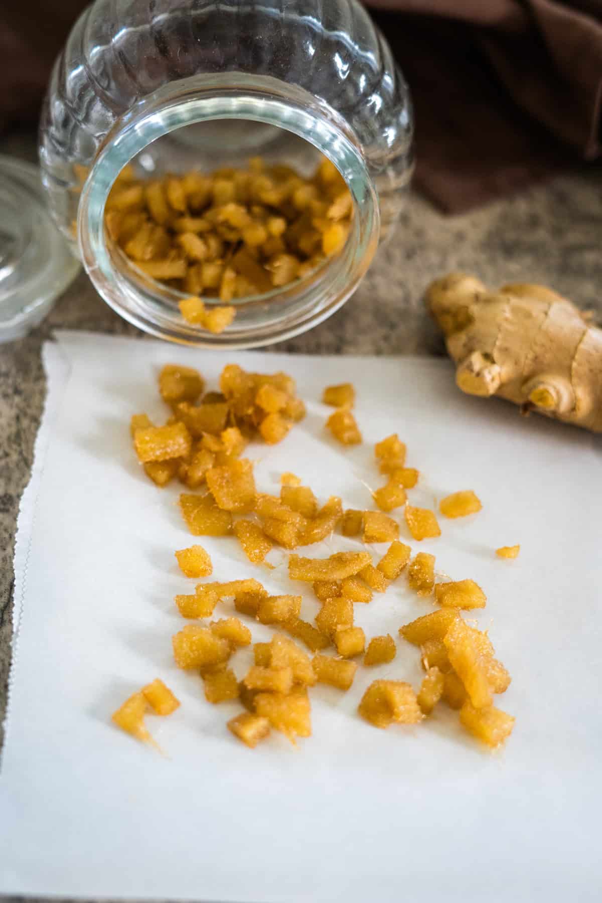 Sugar-free candied ginger pieces spilled from a glass jar onto parchment paper beside a piece of fresh ginger.