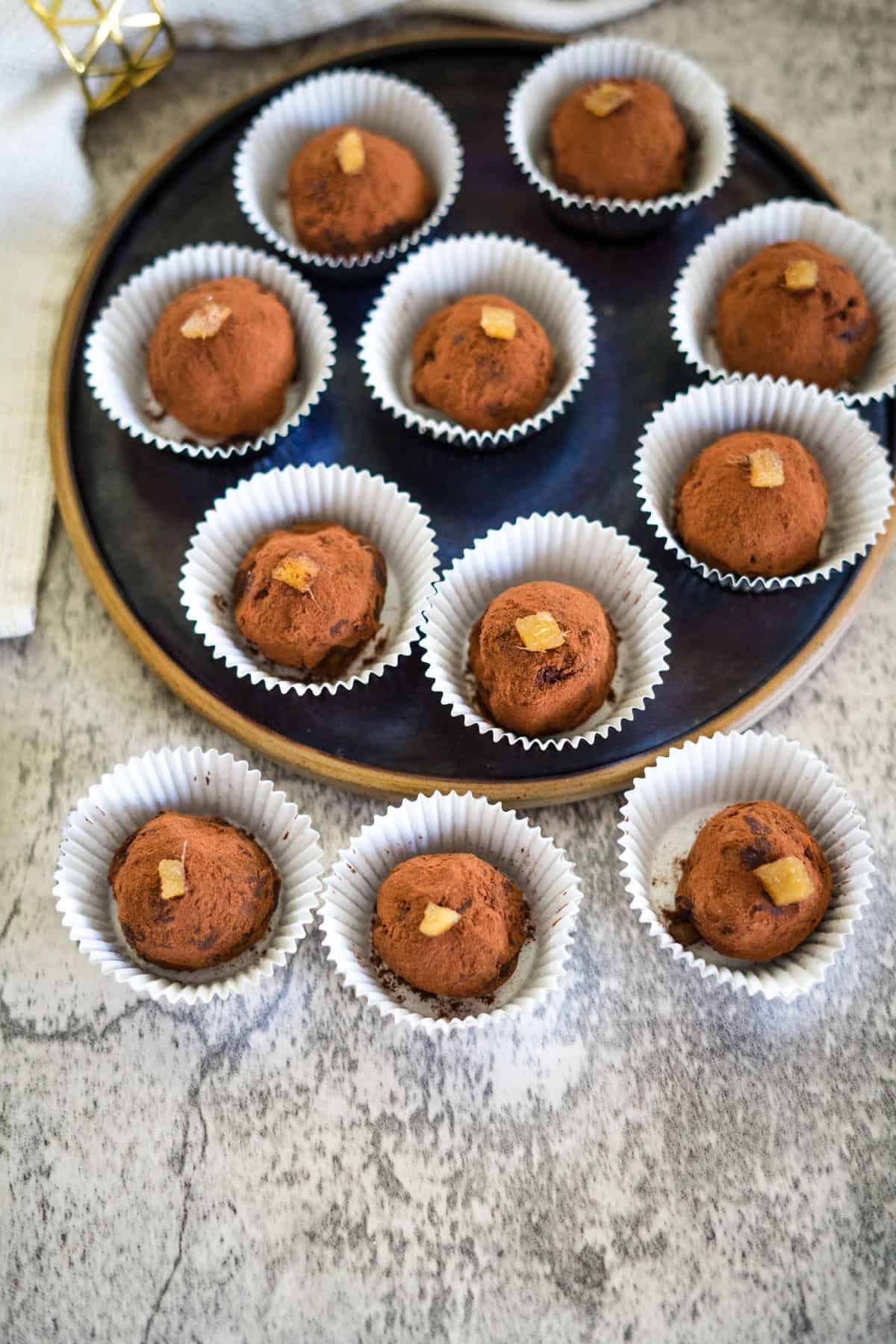 Chocolate truffles with a dusting of cocoa and candied ginger on top, arranged on a round tray with some on a table.