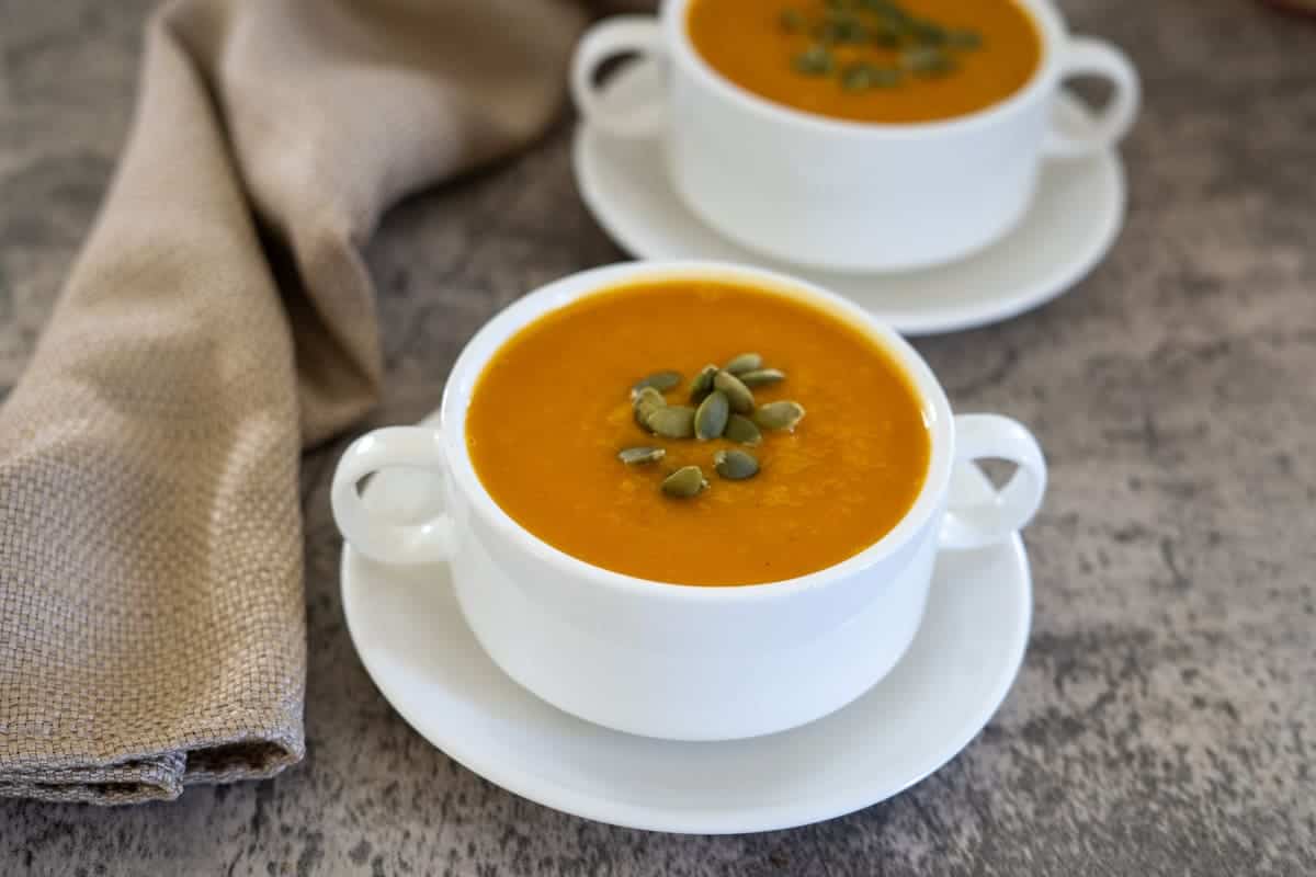 Two white bowls of butternut squash and carrot soup topped with pumpkin seeds rest on a gray surface, accompanied by a folded brown cloth napkin.