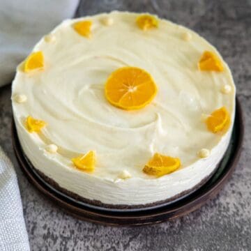 A round orange cake with white frosting, topped with a central orange slice and surrounded by small orange wedge pieces and cream dollops on a dark plate.