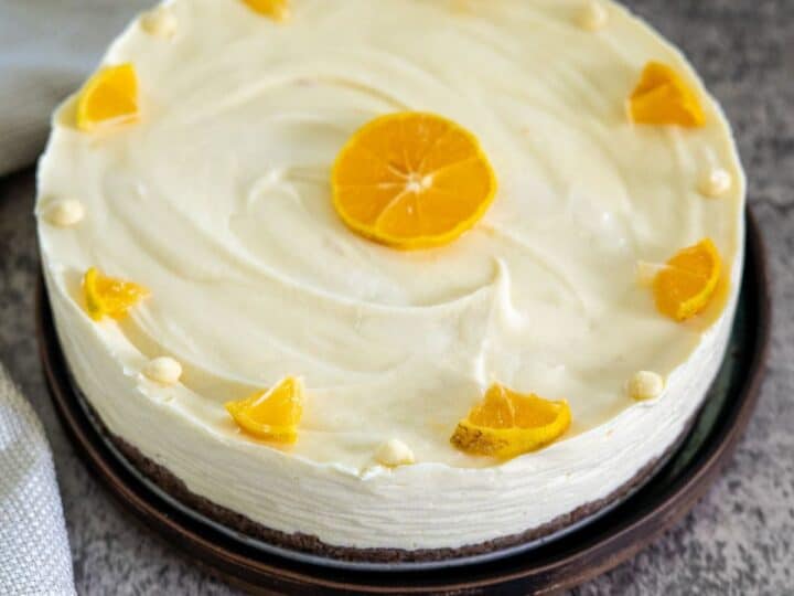 A round orange cake with white frosting, topped with a central orange slice and surrounded by small orange wedge pieces and cream dollops on a dark plate.