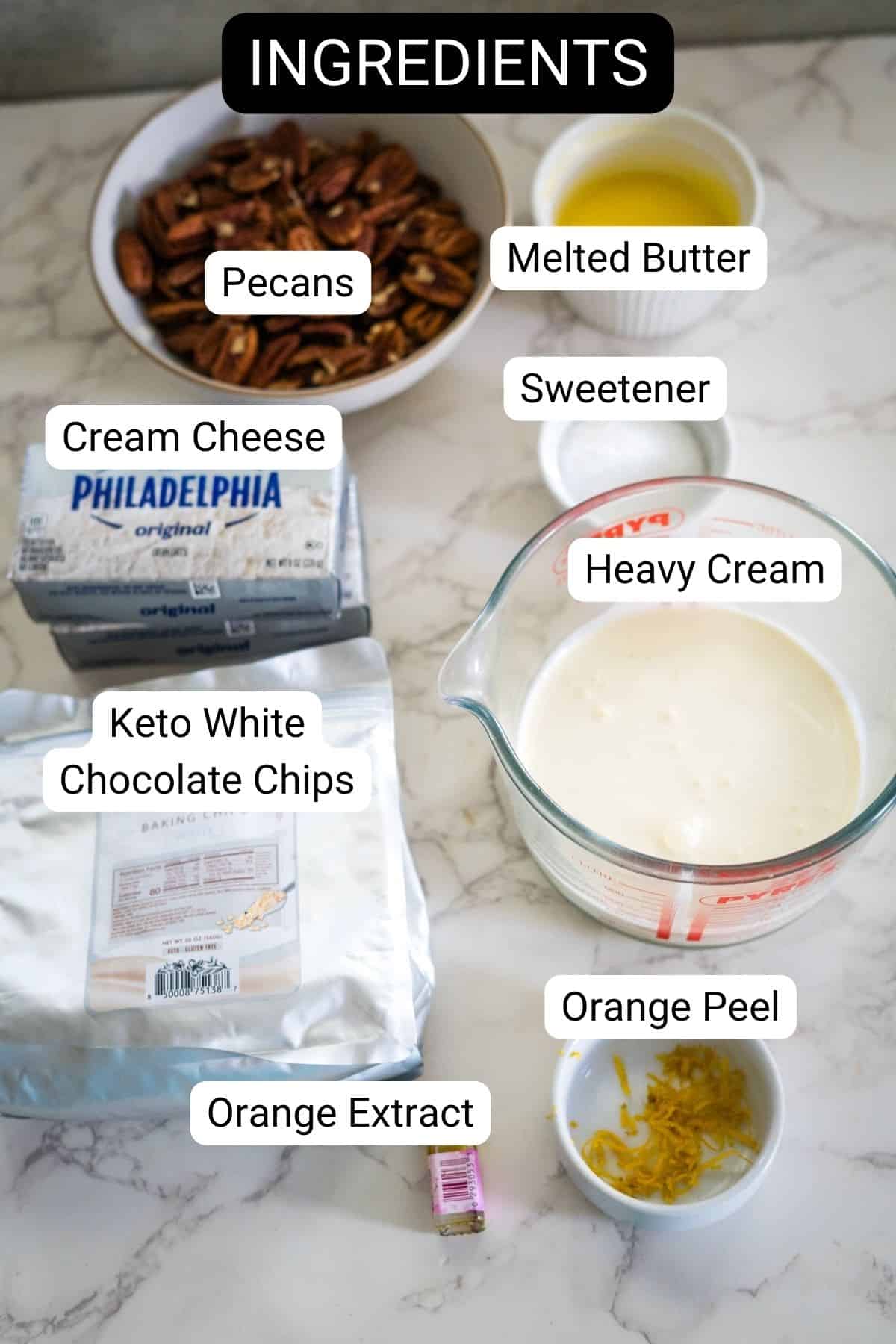 Ingredients on a marble surface include pecans, melted butter, sweetener, cream cheese, heavy cream, keto white chocolate chips, orange peel, and orange extract.