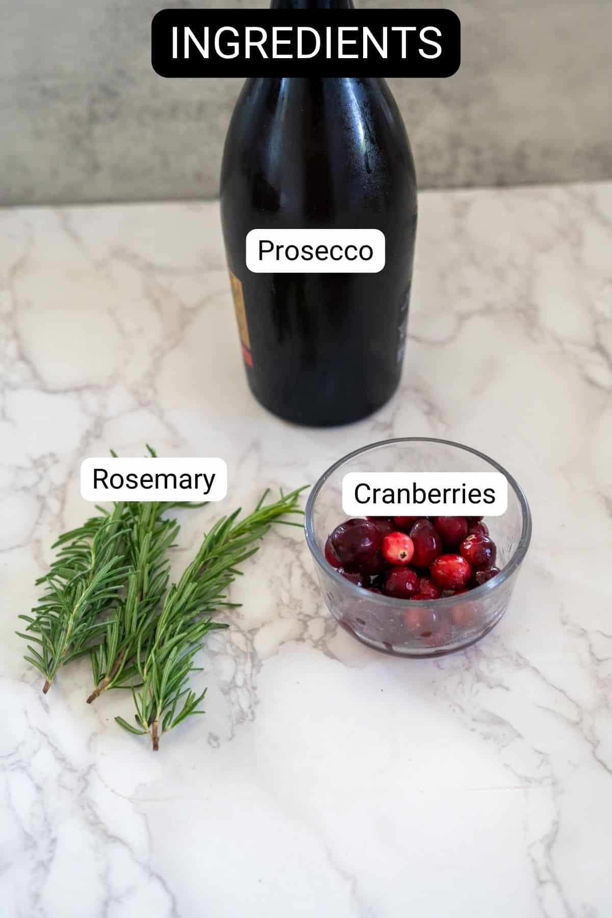 A bottle of prosecco, sprigs of rosemary, and a bowl of cranberries are artfully arranged on a marble surface, evoking the enchanting essence of a snow globe cocktail.