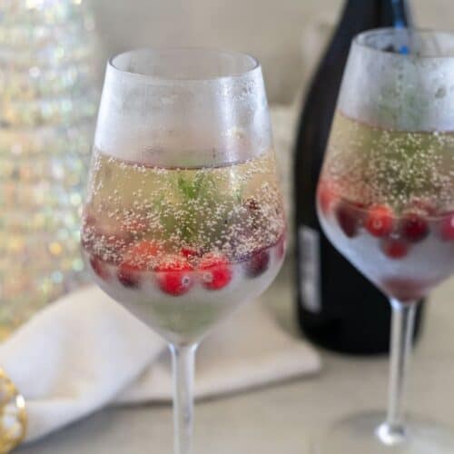 Two wine glasses filled with a chilled fizzy drink, garnished with red berries and green leaves. A bottle is in the background.