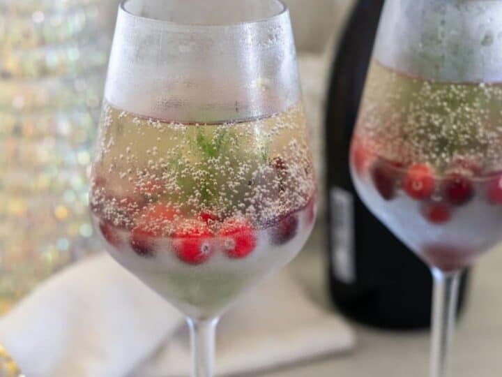 Two wine glasses filled with a chilled fizzy drink, garnished with red berries and green leaves. A bottle is in the background.