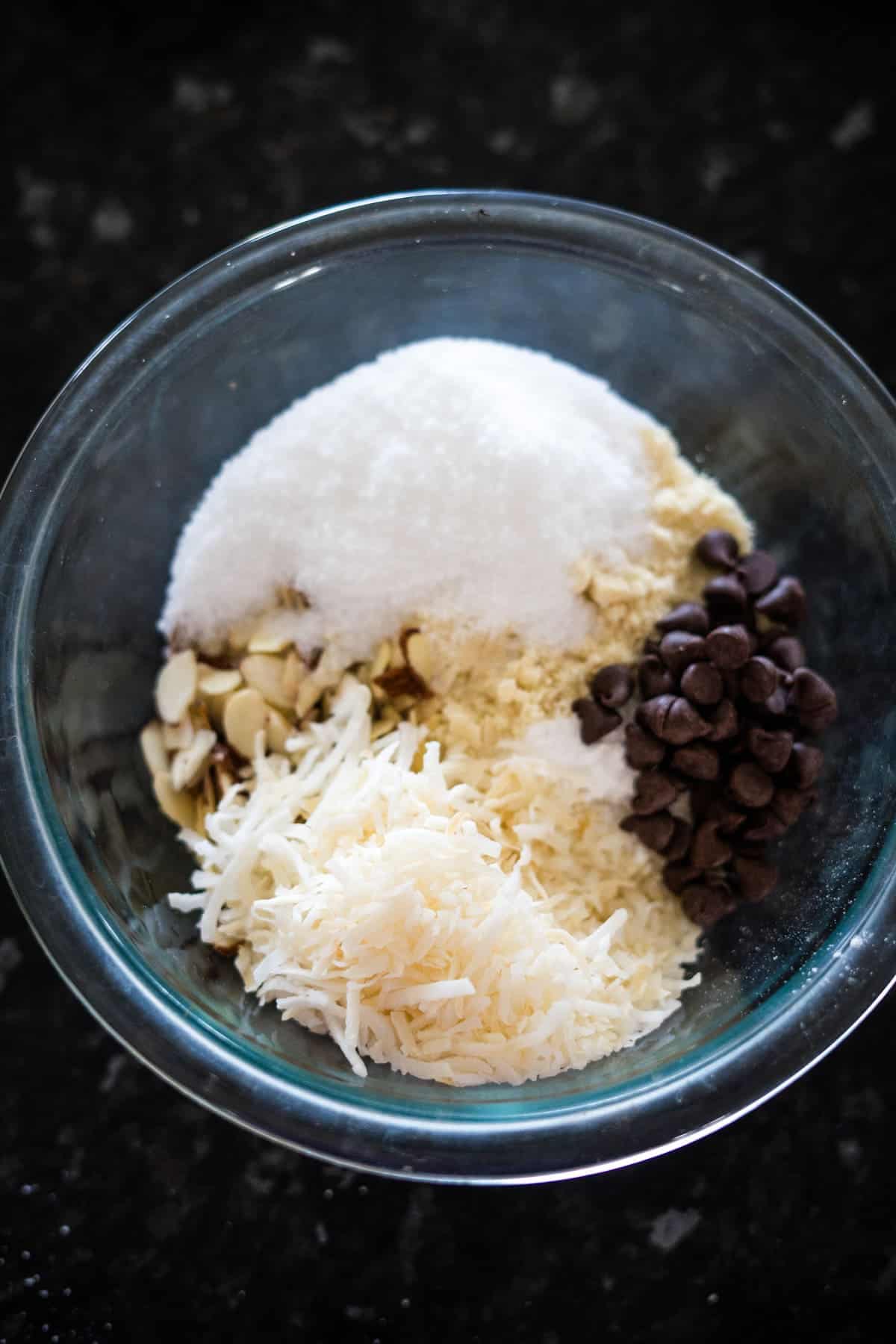 A glass bowl containing shredded coconut, chocolate chips, sliced almonds, and sugar, placed on a dark surface.