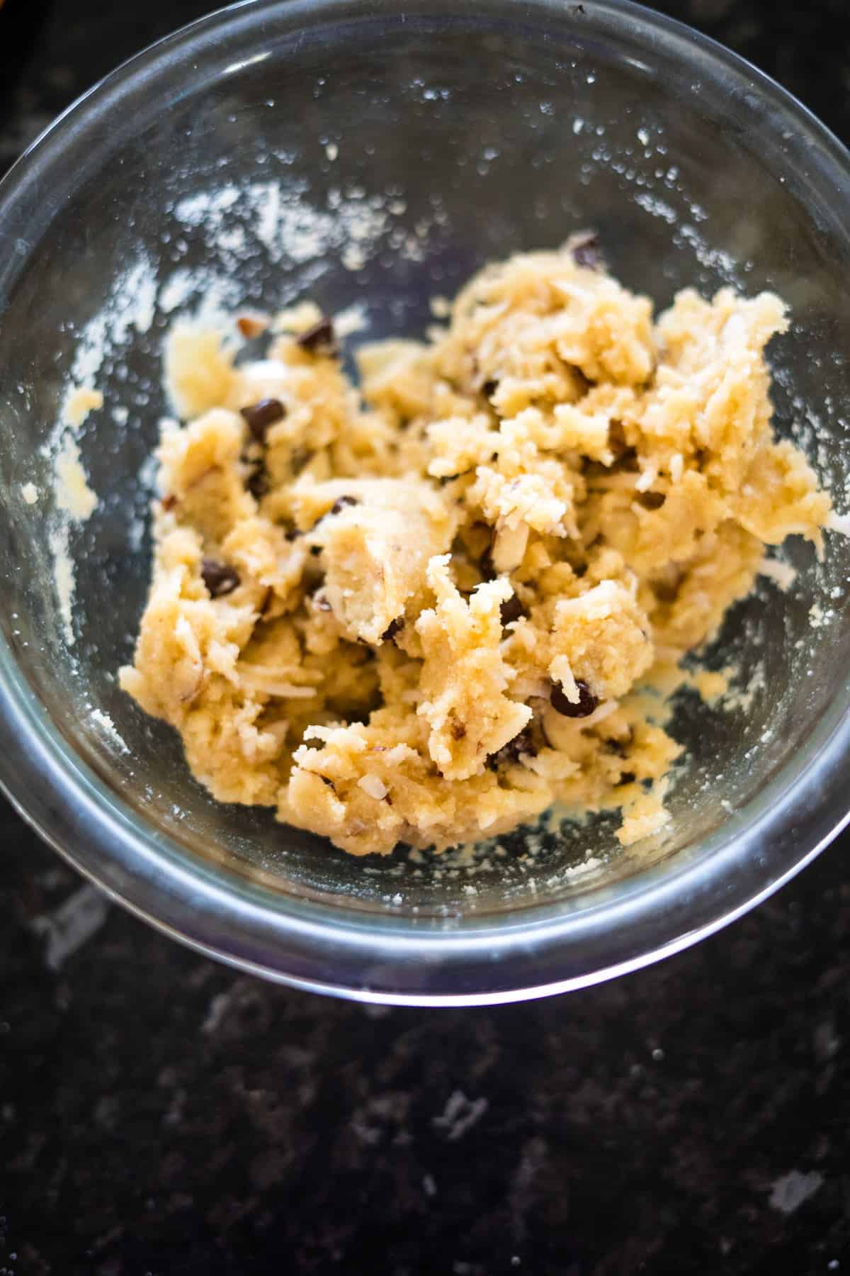 A glass bowl containing chunky cookie dough with visible chocolate chips on a dark countertop.