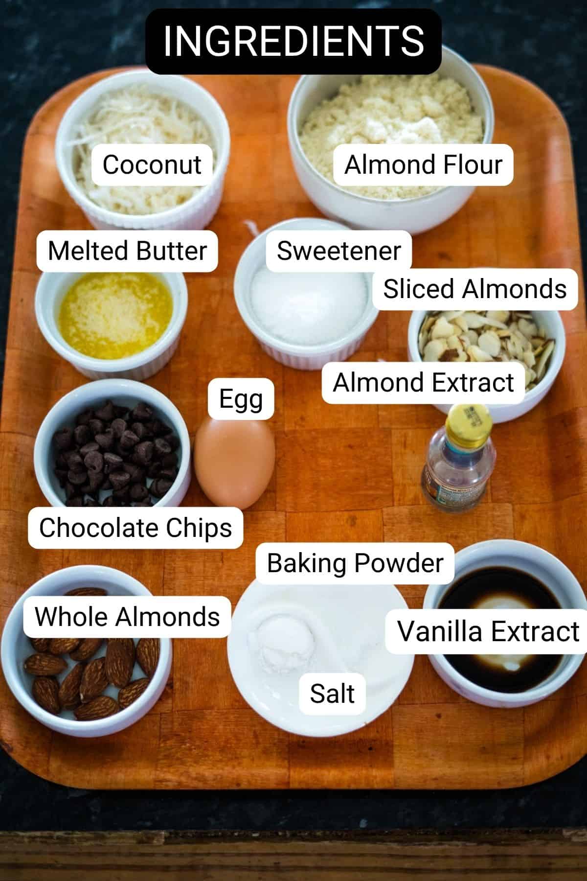 Wooden tray with labeled ingredients: coconut, almond flour, melted butter, sweetener, sliced almonds, egg, almond extract, chocolate chips, whole almonds, baking powder, salt, and vanilla extract.