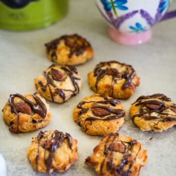 Seven almond cookies topped with chocolate drizzle are arranged on a gray surface. A floral teacup and green kettle are in the background.