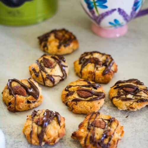 Seven almond cookies topped with chocolate drizzle are arranged on a gray surface. A floral teacup and green kettle are in the background.