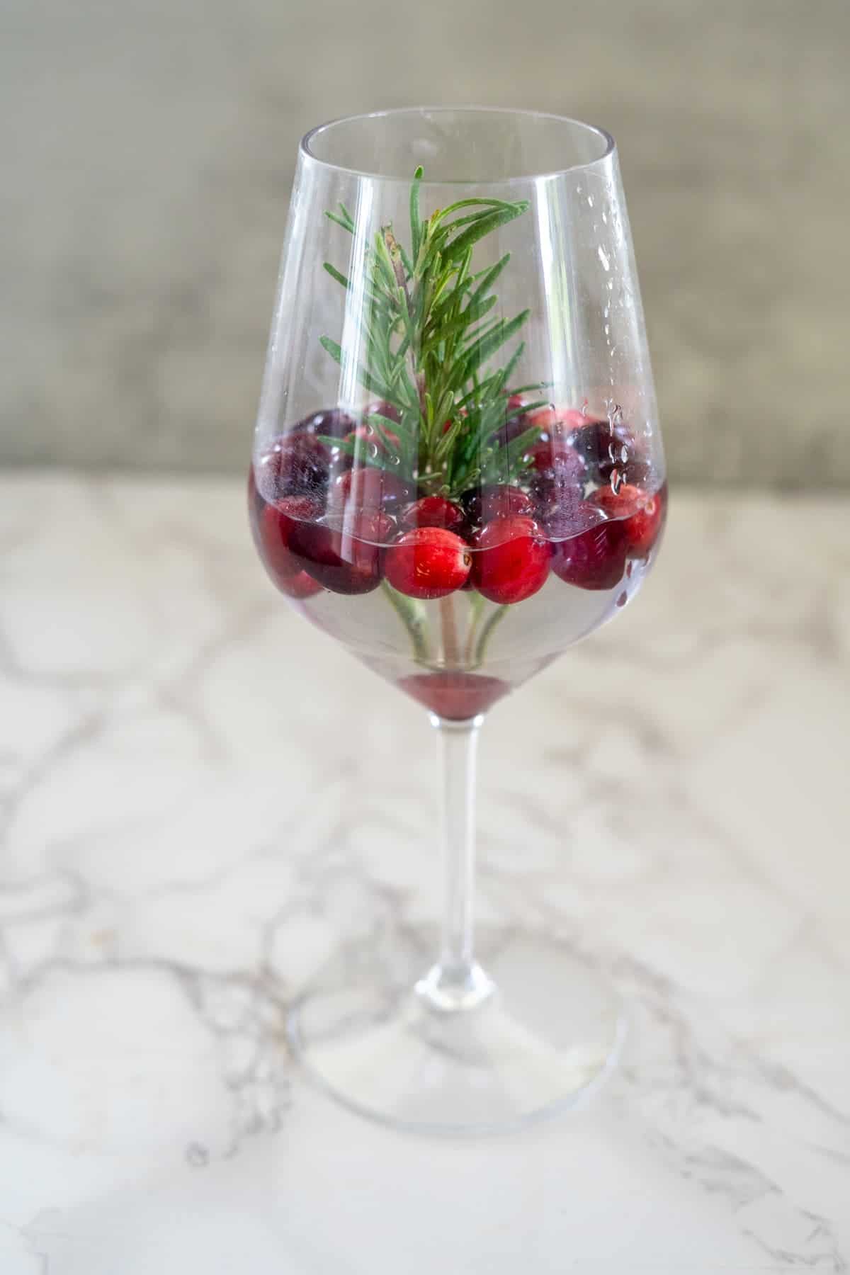 A wine glass filled with clear liquid, cranberries, ice cubes, and a sprig of rosemary creates a picturesque snow globe cocktail.