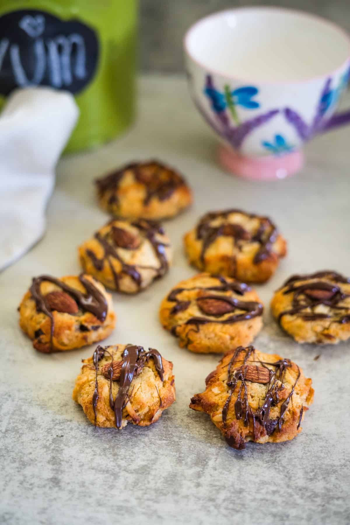 Keto almond joy cookies topped with almonds and drizzled with chocolate are arranged on a table. A tea cup and a jar sit in the background.