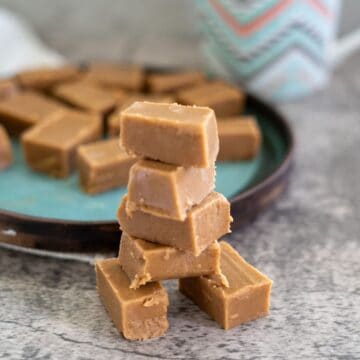 A stack of brown fudge pieces is in focus, with more pieces on a blue plate in the background. A patterned mug is partially visible.