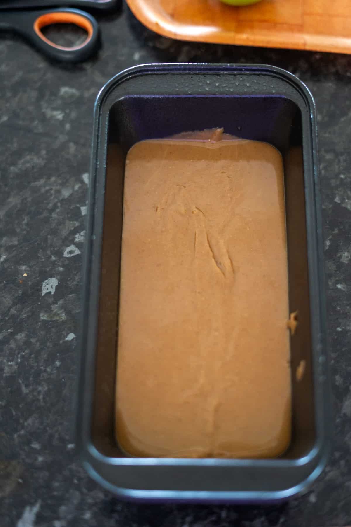 A loaf pan filled with smooth brown keto peanut butter fudge batter rests on a dark countertop. Scissors and a wooden board are visible in the background.